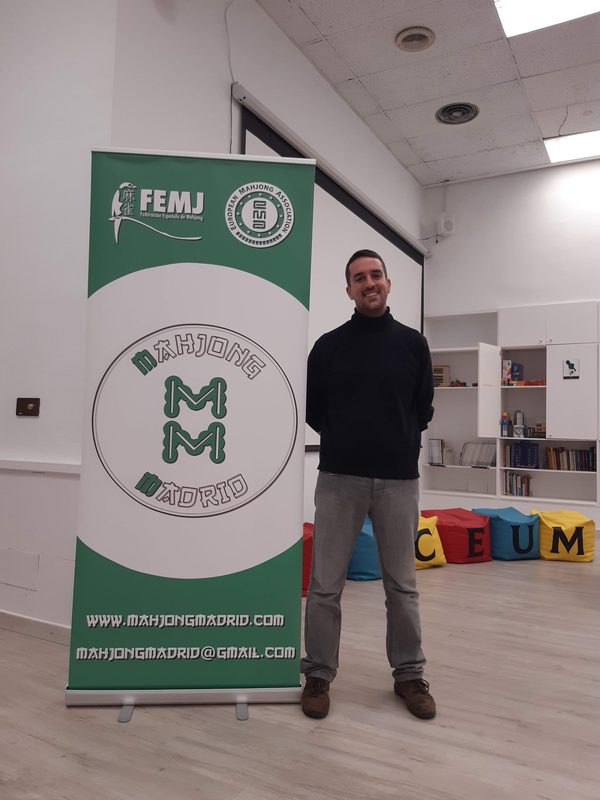 Ernesto sonriendo junto al cartel de Mahjong Madrid, la FEMJ y la EMA